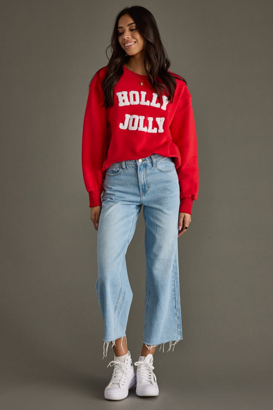 Woman wearing a red 'Holly Jolly' sweater with light blue jeans and white sneakers on a gray background