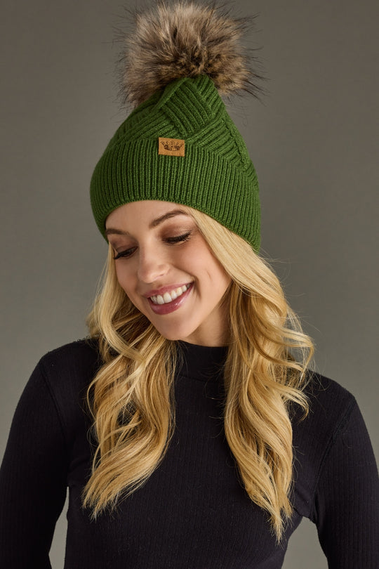 Woman wearing a green knit beanie with a fur pom-pom on a gray background