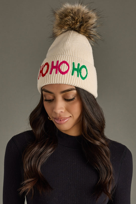 A woman models the Panache Apparel Collection’s Ho Ho Ho Pom Hat, featuring a beige knit, faux fur pom, and red, pink, and green “HO HO HO” lettering, paired with a black ribbed sweater against a gray background.