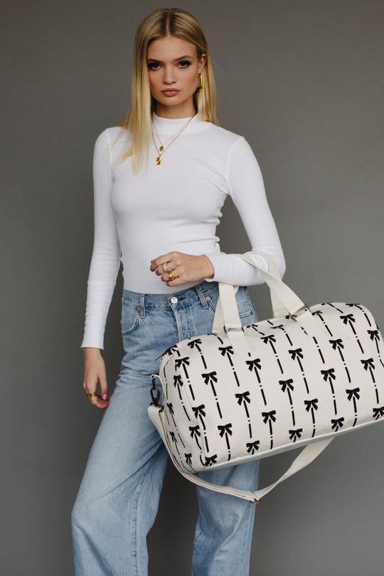 Woman holding a white duffel bag with black bow pattern against a gray background