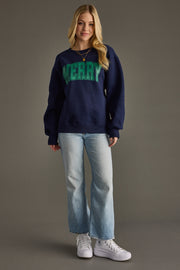 Person wearing a navy sweatshirt with 'MERRY in green, light blue jeans, and white sneakers on a gray background