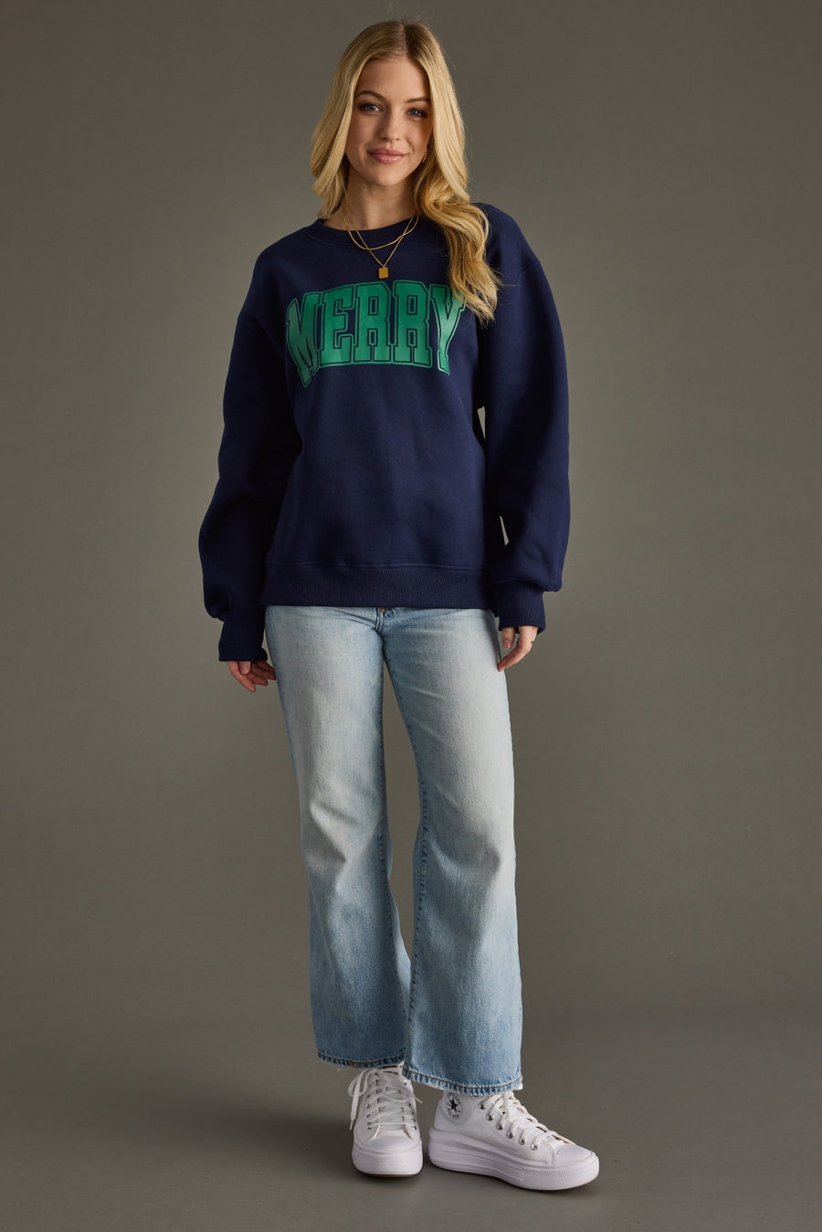 Person wearing a navy sweatshirt with 'MERRY in green, light blue jeans, and white sneakers on a gray background