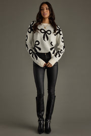 Woman wearing a white sweater with black bow patterns, black pants, and black boots on a gray background