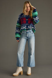 A woman with long blonde hair poses against a plain background in the Holly Cardigan from Panache Apparel Collection, paired with light blue high-waisted jeans and beige heeled boots. Her left hand is lifted to her head.