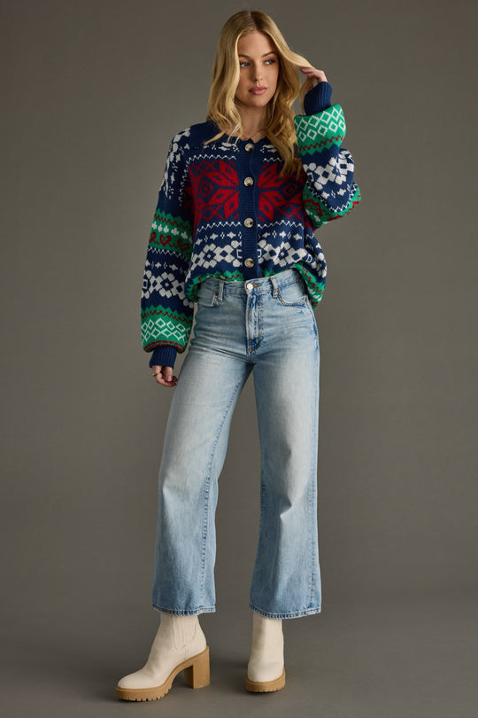 A woman with long blonde hair poses against a plain background in the Holly Cardigan from Panache Apparel Collection, paired with light blue high-waisted jeans and beige heeled boots. Her left hand is lifted to her head.