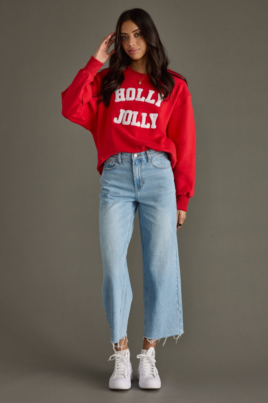 A woman stands against a plain background in the Panache Apparel Collection Holly Jolly Sweatshirt - Red, paired with light blue high-waisted jeans with frayed hems and white sneakers. She touches her hair and smiles slightly.