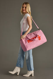 Woman holding a pink duffel bag with 'Merry' written on it against a gray background
