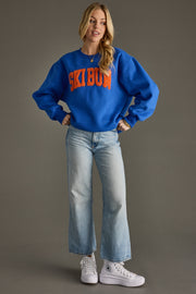 Person wearing a blue sweatshirt with 'Ski Bum' text, light blue jeans, and white sneakers on a gray background