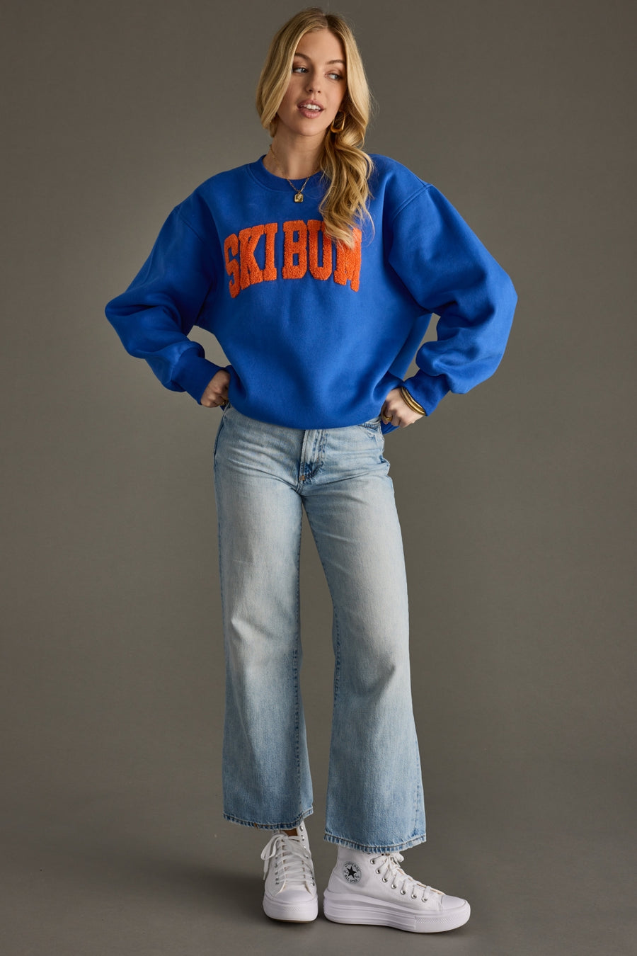 Person wearing a blue sweatshirt with 'Ski Bum' text, light blue jeans, and white sneakers on a gray background