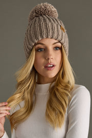Woman wearing a taupe knitted beanie with a pom-pom against a gray background