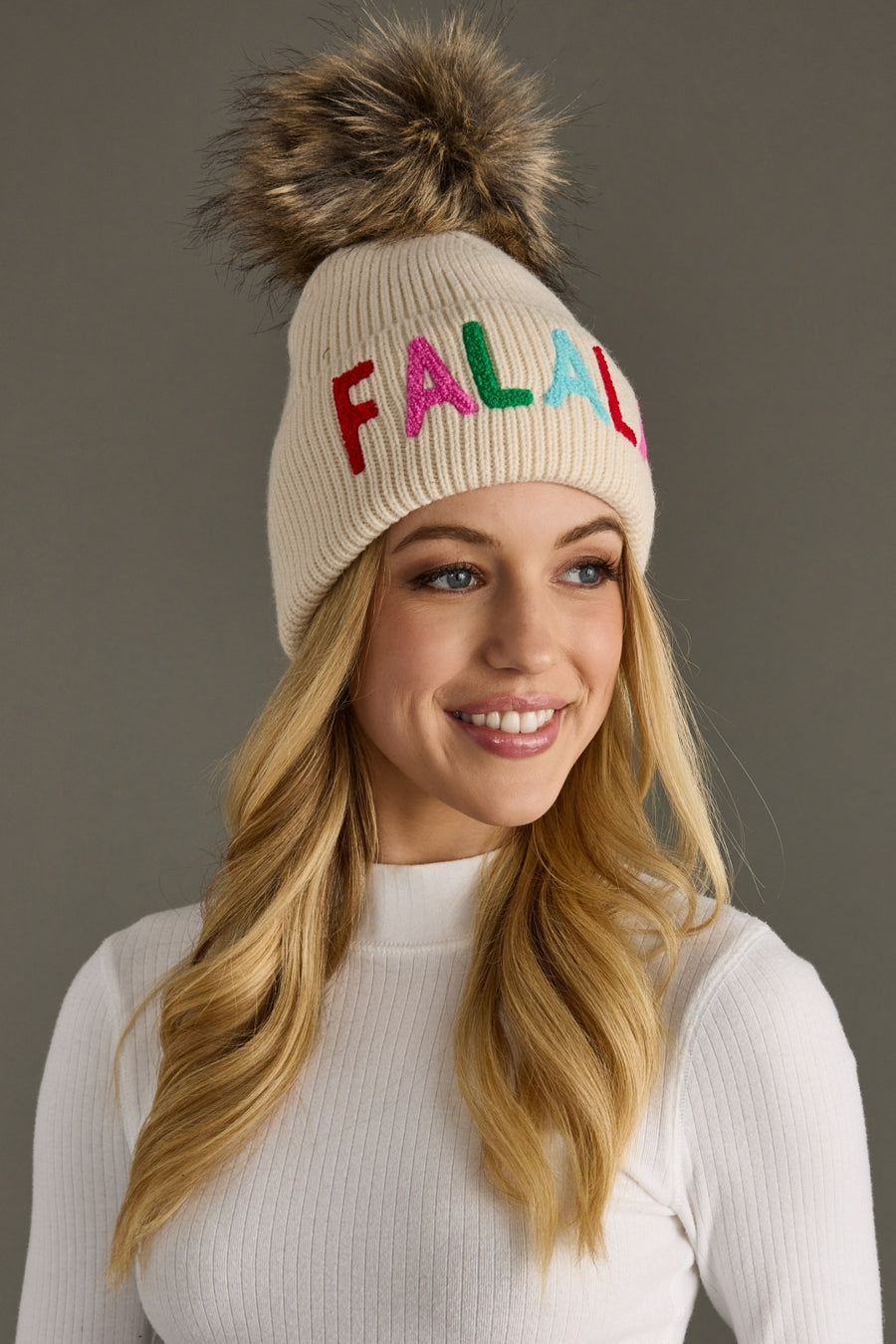 Woman wearing a beige knit beanie with colorful letters on a gray background