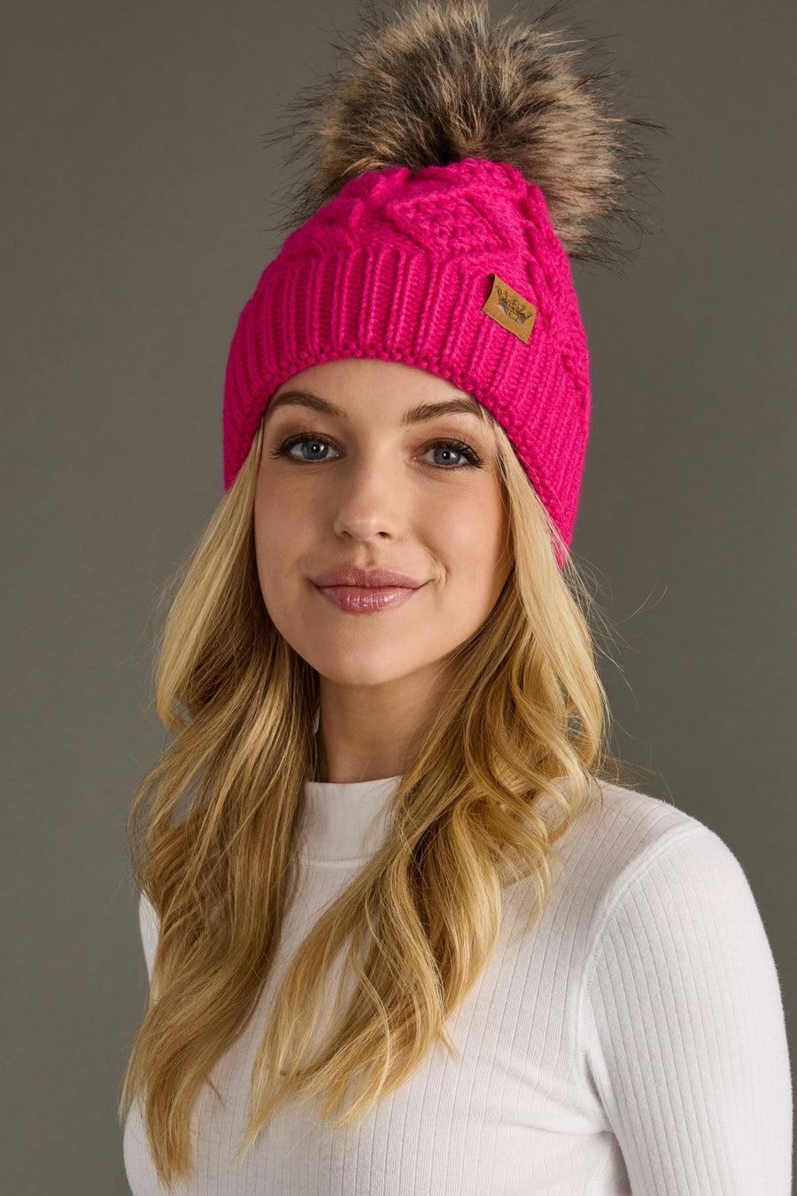 Woman wearing a bright pink knit beanie with a fur pom-pom on a gray background
