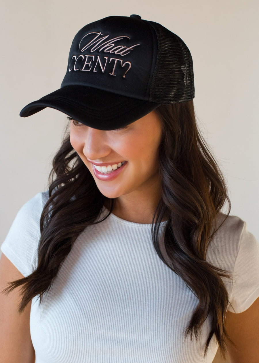 She’s wearing a black trucker hat with the phrase “What ACCENT?” embroidered across the front in a mix of script and block lettering. Paired with loose waves and a simple white tee, the vibe is playful, confident, and a little sassy in the best way.
