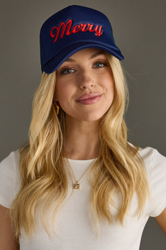 Woman wearing a navy trucker hat with 'Merry' in red text on a gray background