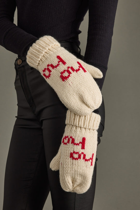 Pair of cream mittens with red text held by a person wearing black gloves against a gray background