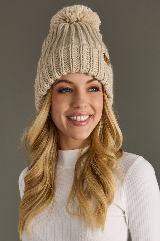 Woman wearing a beige knitted beanie with a pom-pom against a gray background