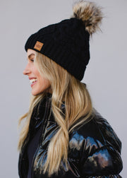 Woman wearing a black knit beanie with a pom-pom on a plain background