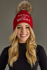 Person wearing a red beanie with 'Merry and Bright' text and a fur pom-pom on a gray background