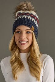 A smiling woman with long blonde hair wears the Ezra Pom Hat by Panache Apparel Collection, a fleece-lined knit hat with white and red patterns and a faux fur pom-pom, plus a white ribbed turtleneck sweater, against a plain gray background.