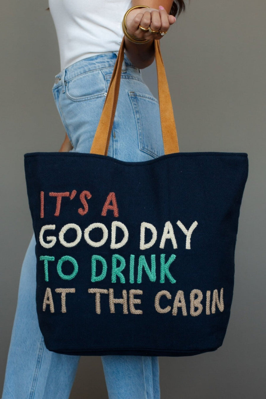 A person in blue jeans holds the Panache Apparel Collection Drink At The Cabin Tote, a navy cabin bag featuring tan leather suede straps and colorful lettering that says, "ITS A GOOD DAY TO DRINK AT THE CABIN.