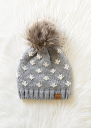Gray knit beanie with white floral patterns and a fluffy pom-pom on a white background