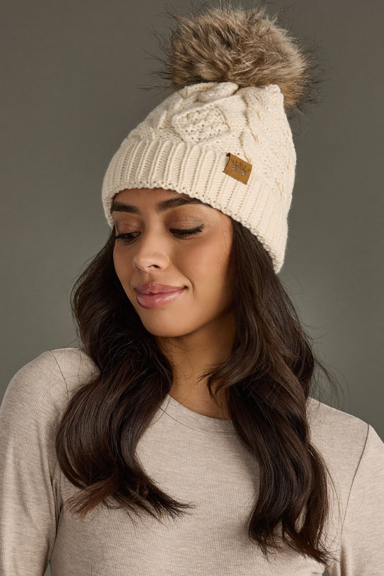 Woman wearing a cream knit beanie with a pom-pom against a gray background