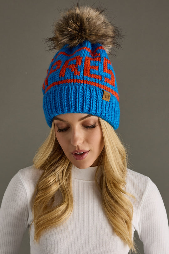 Blue knit beanie with orange text and fur pom-pom worn by a person against a gray background