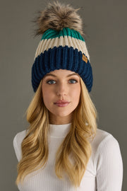 A woman with long blonde hair models the Panache Apparel Collection So Cozy Pom Hat, a blue, green, and cream ribbed knit beanie with a large faux fur pom-pom, paired with a white ribbed sweater against a gray background.