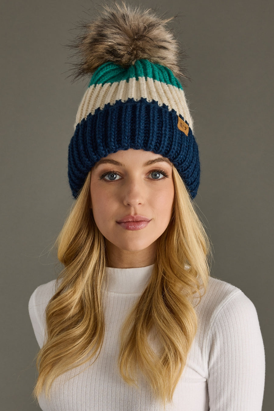 A woman with long blonde hair models the Panache Apparel Collection So Cozy Pom Hat, a blue, green, and cream ribbed knit beanie with a large faux fur pom-pom, paired with a white ribbed sweater against a gray background.