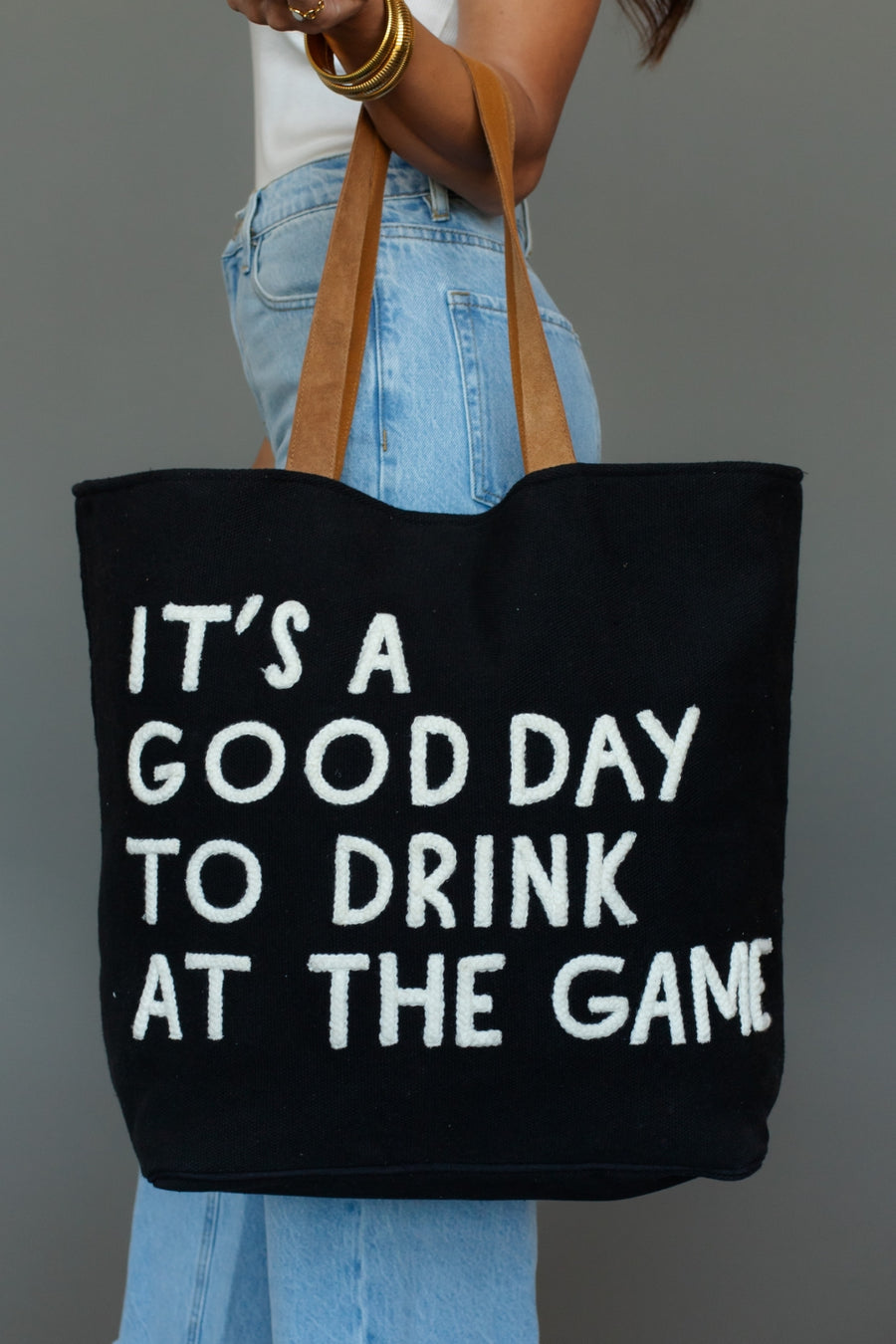 Black tote bag with white text 'It's a good day to drink at the game' held by a person wearing blue jeans.