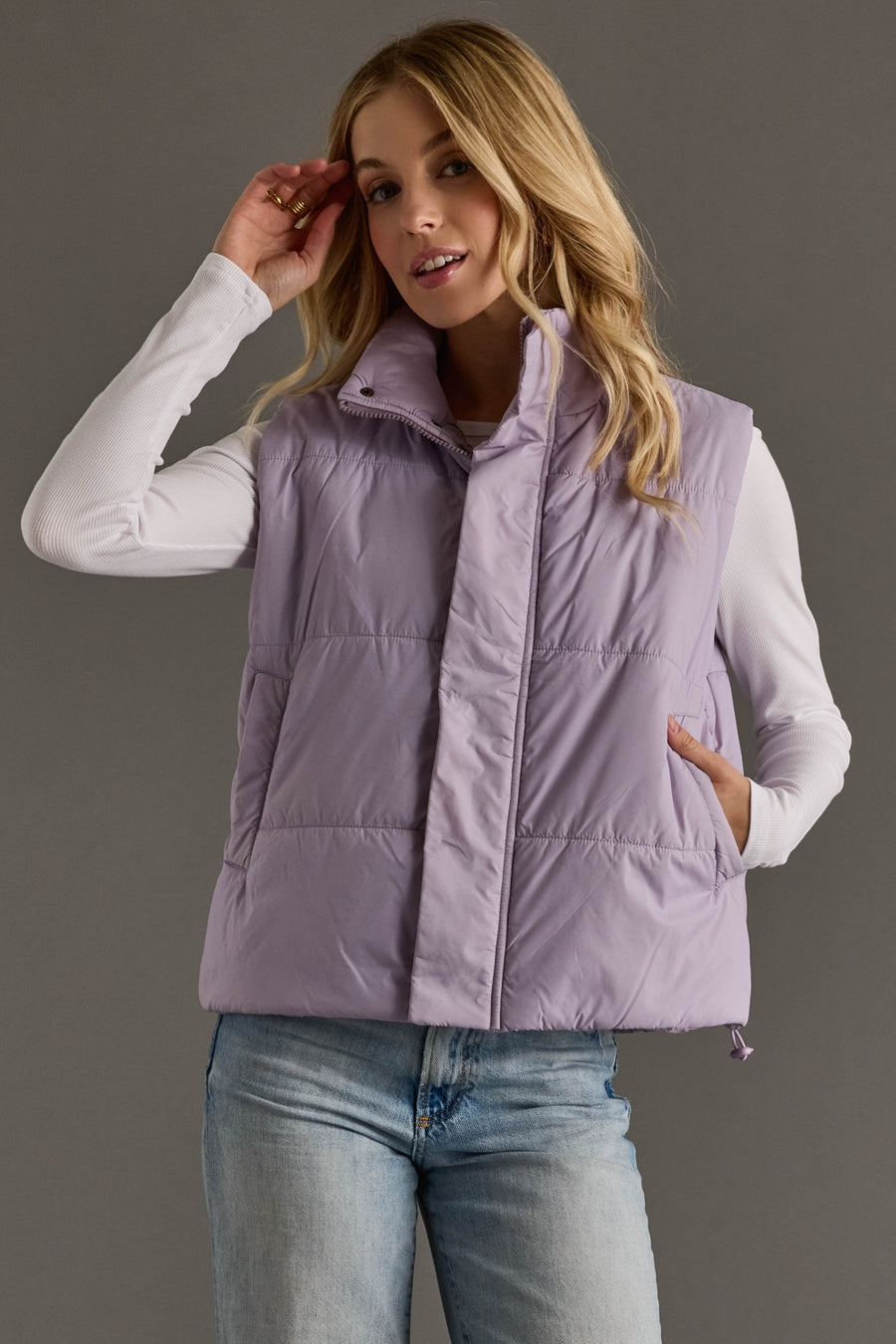Woman wearing a light purple puffer vest over a white shirt and blue jeans on a gray background