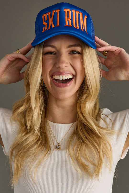 Woman wearing a blue cap with 'SKI BUM' text, smiling against a gray background