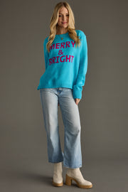Woman wearing a blue sweater with 'Merry & Bright' text, light blue jeans, and beige boots on a gray background