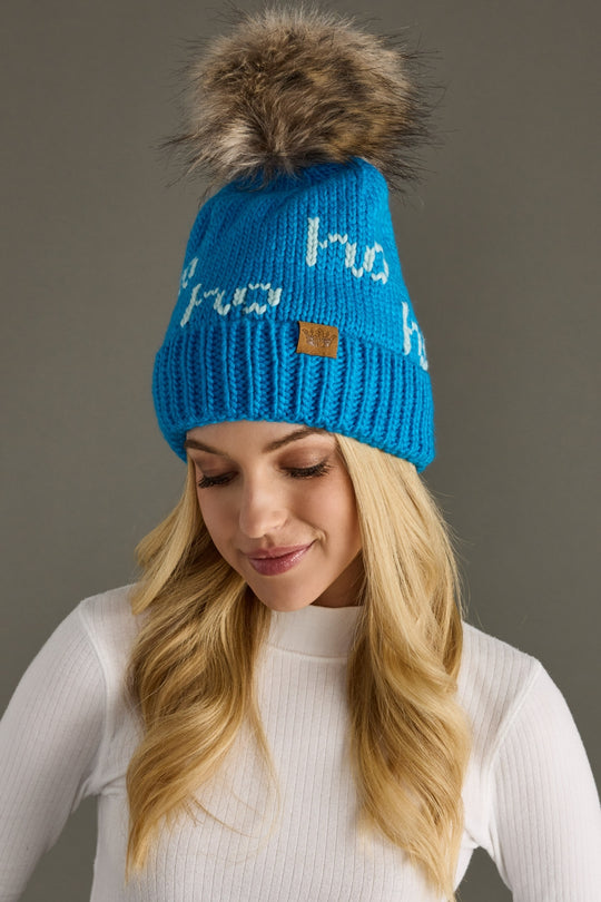 Blue knit beanie with fur pom-pom worn by a person against a gray background
