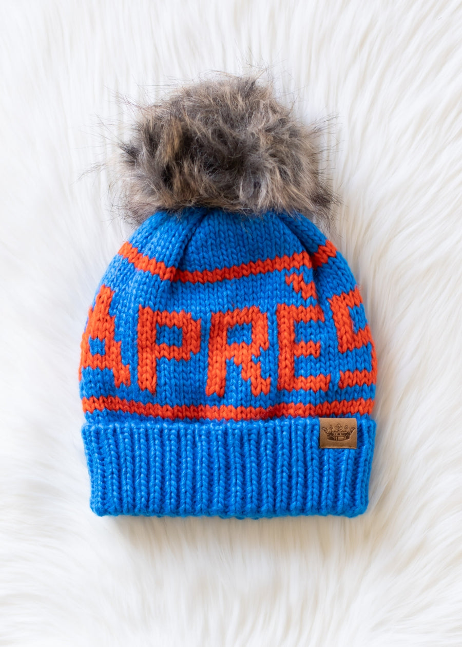 Blue knit beanie with orange text and fur pom-pom on a white surface