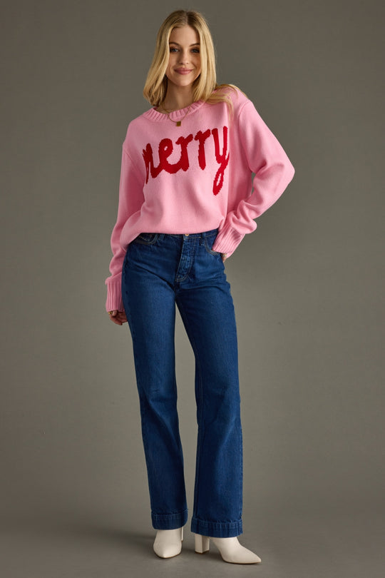 Woman wearing a pink sweater with 'Merry' text and blue jeans on a gray background