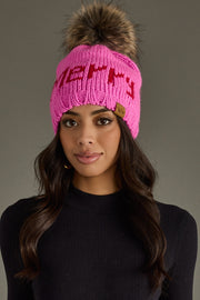 Woman wearing a pink knit beanie with a fur pom-pom on a gray background