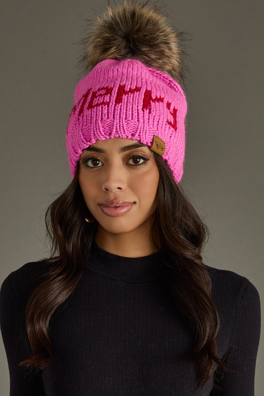 Woman wearing a pink knit beanie with a fur pom-pom on a gray background
