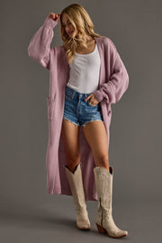 Woman wearing a long pink cardigan, white top, denim shorts, and beige cowboy boots on a gray background