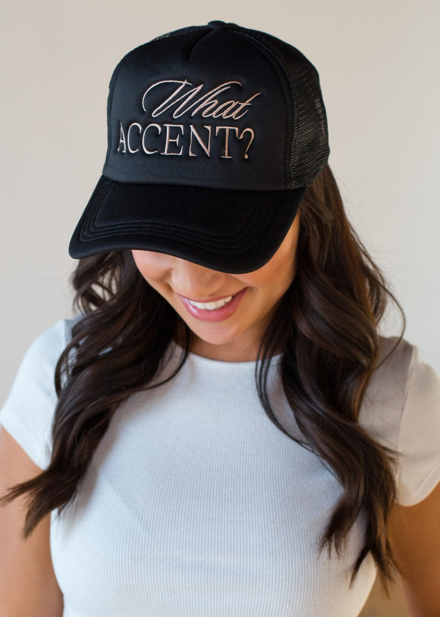 She’s wearing a black trucker hat with the phrase “What ACCENT?” embroidered across the front in a mix of script and block lettering. Paired with loose waves and a simple white tee, the vibe is playful, confident, and a little sassy in the best way.
