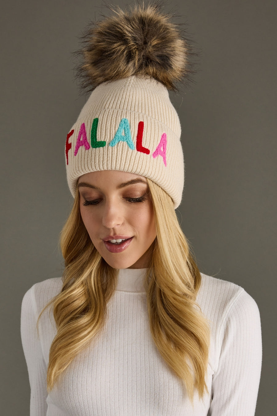Beige knit hat with colorful 'FALALA' text and fur pom-pom worn by a model against a gray background