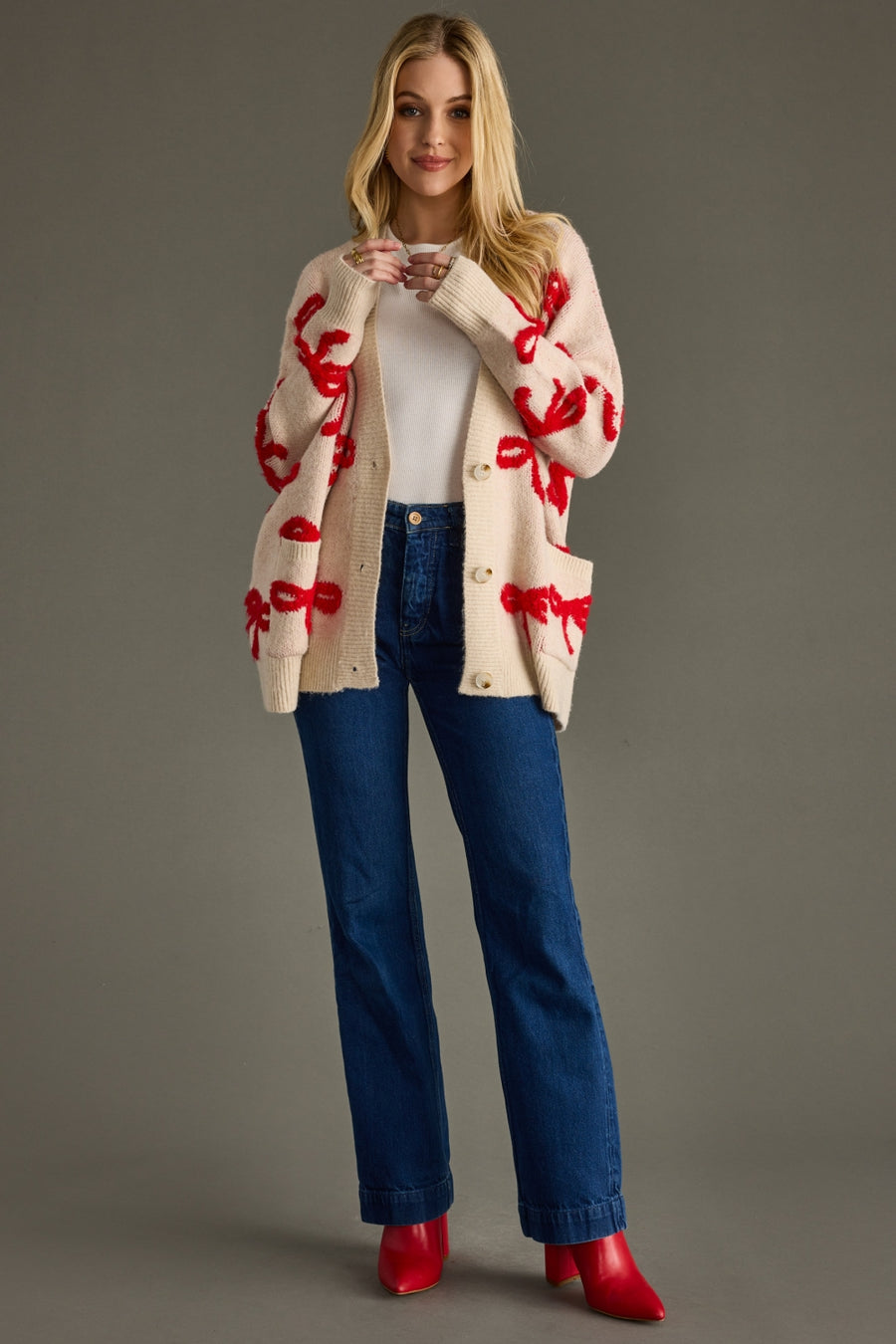 Woman wearing a cream cardigan with red patterns, white shirt, blue jeans, and red shoes on a gray background