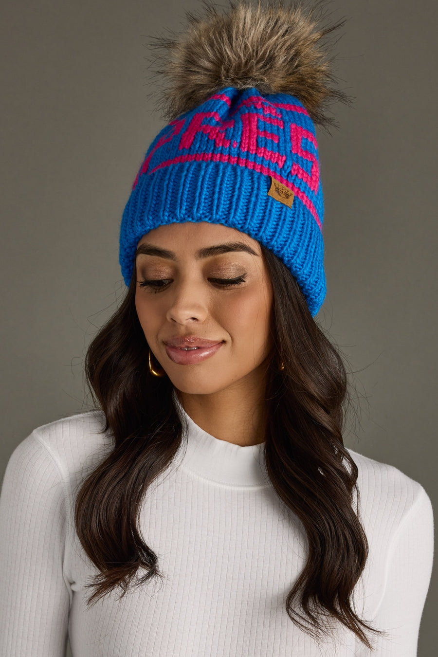 Woman wearing a blue and pink beanie with a fur pom-pom on a gray background