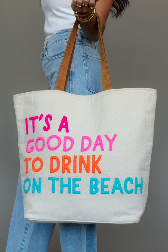 Tote bag with colorful text 'It's a good day to drink on the beach' held by a person.