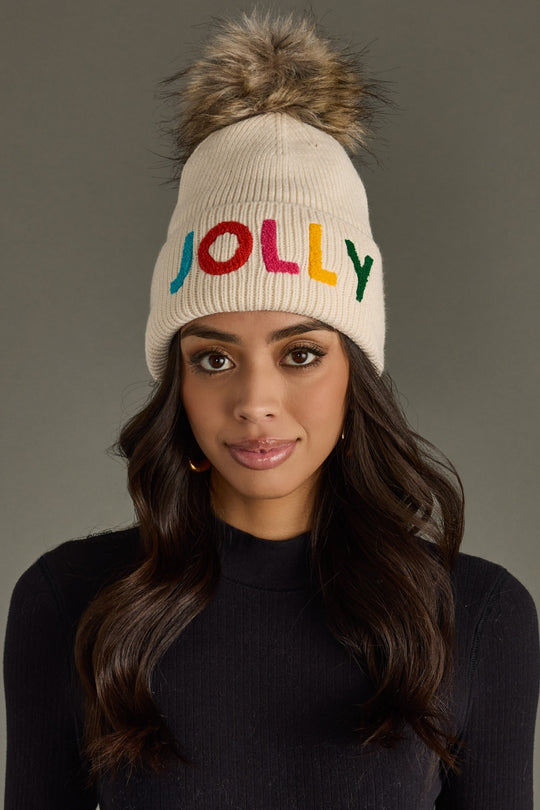 Woman wearing a beige knit beanie with colorful 'JOLLY' text and fur pom-pom on a gray background