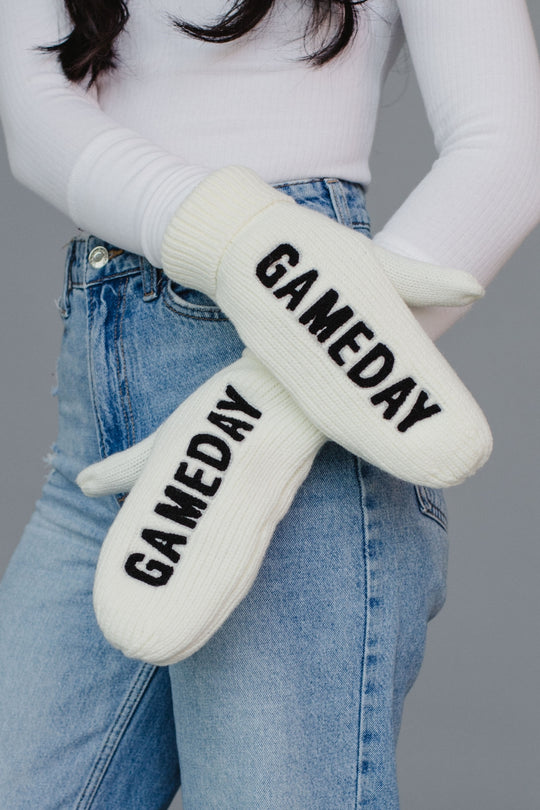 White mittens with 'GAMEDAY' text worn by a person wearing jeans and a white long-sleeve shirt.