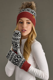 A knit beanie features a classic Fair Isle geometric pattern in cream and black with a solid rust-red ribbed cuff and small tan leather logo patch. Topped with a luxurious faux-fur pom-pom, this winter hat delivers Nordic-inspired alpine style with cozy warmth.
