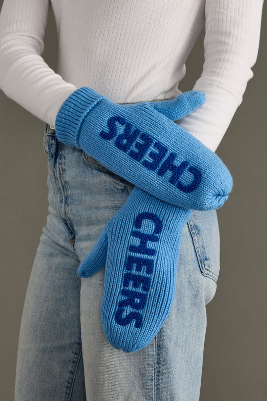 Blue mittens with 'CHEERS' text worn by a person in a white long-sleeve shirt and jeans.