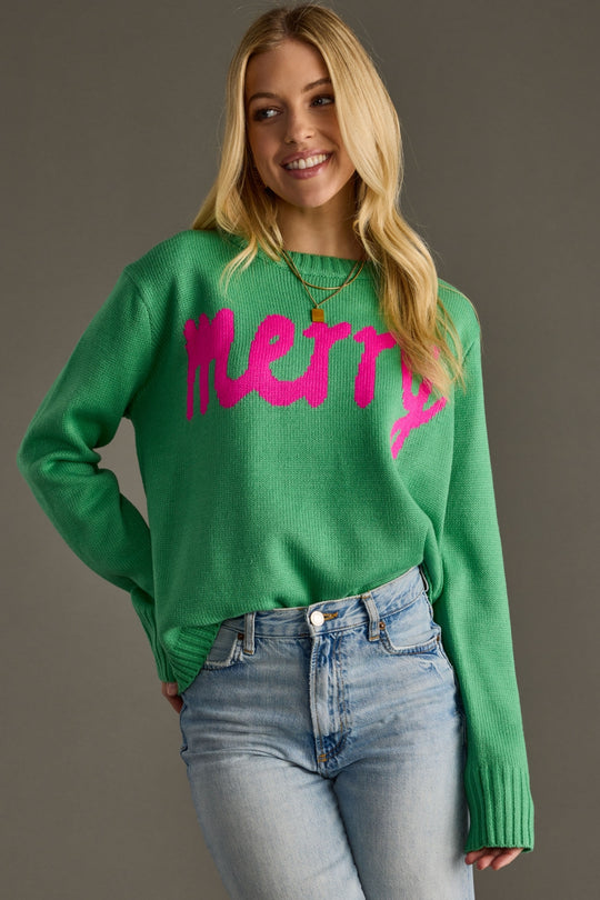Woman wearing a green sweater with pink text and blue jeans against a gray background