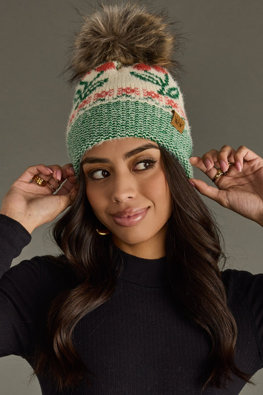 Woman wearing a green and beige knitted beanie with a pom-pom against a gray background
